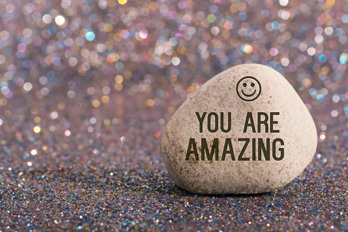 Smooth stone with smiley face reading 'you are amazing' on glitter background